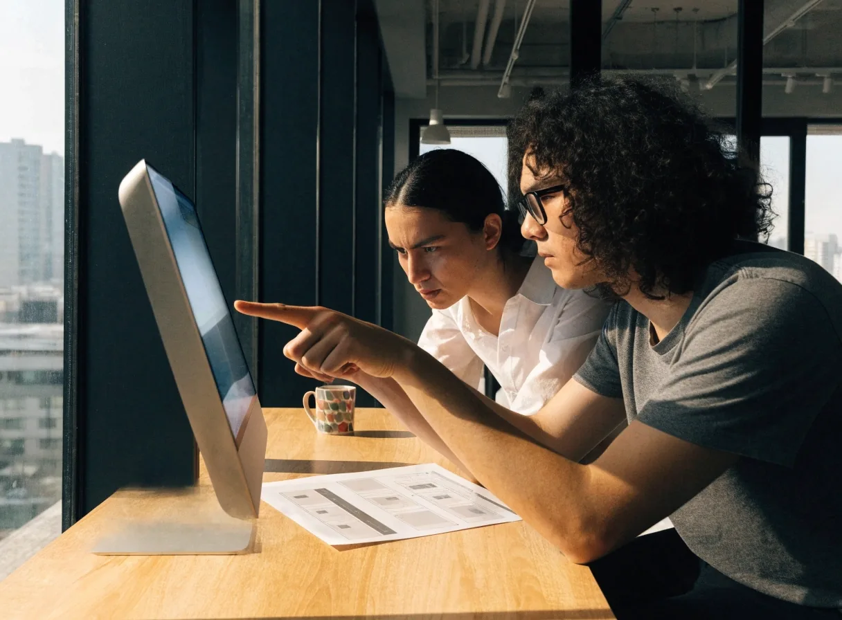 A designer and developer reviewing a printed site map beside a glowing monitor in a Singapore office